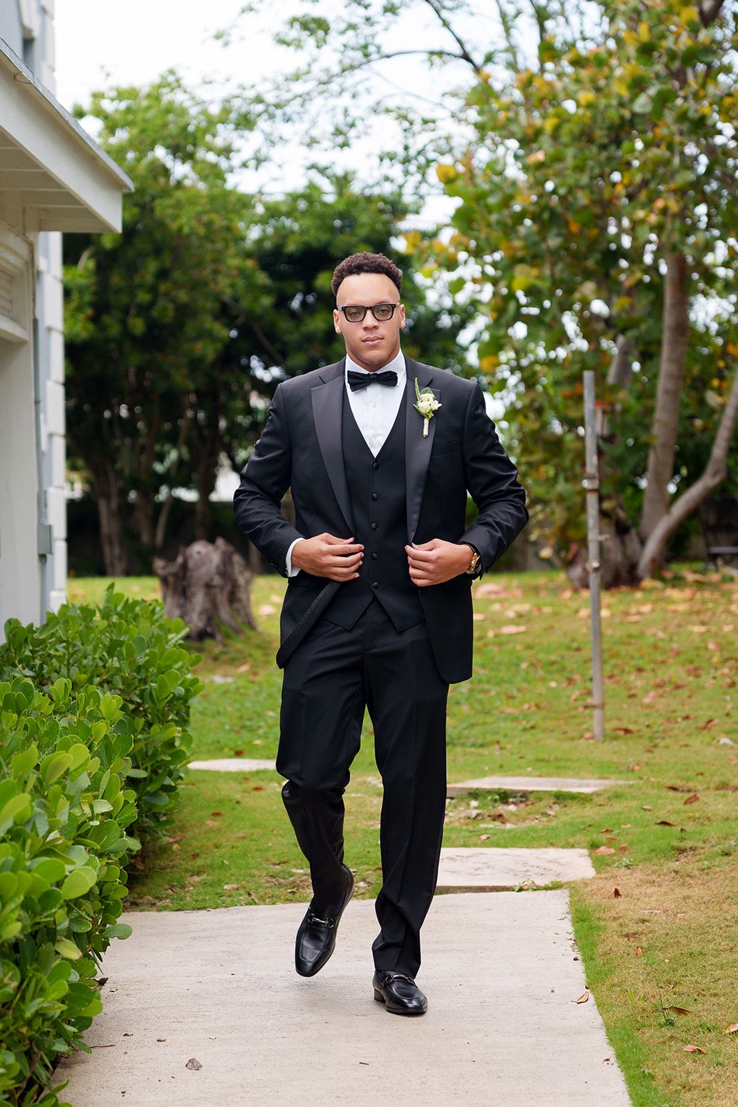 A groom in a black tuxedo with boutonniere and bow tie walks confidently along an outdoor pathway.