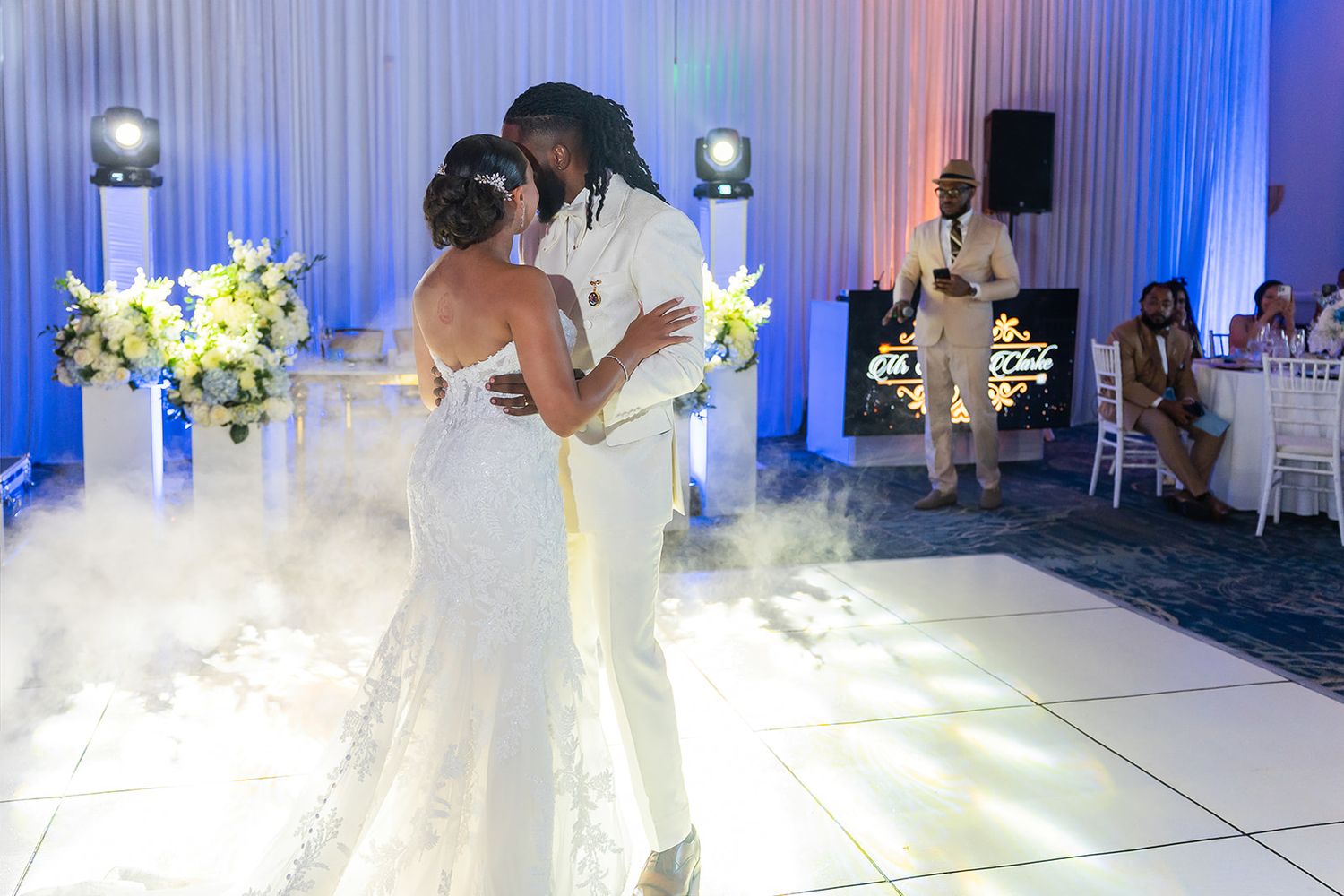 Bride and groom share first dance on white floor surrounded by fog at elegant blue-lit wedding reception.