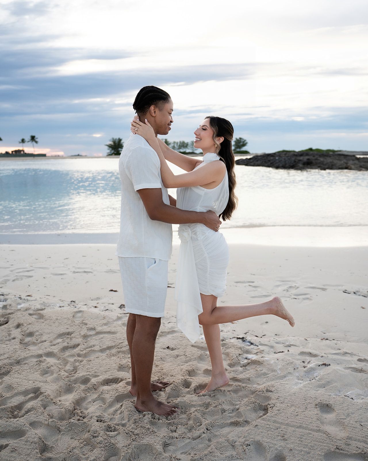 Romantic couple embracing on a tropical beach at sunset, both wearing white outfits, with calm blue waters behind them.