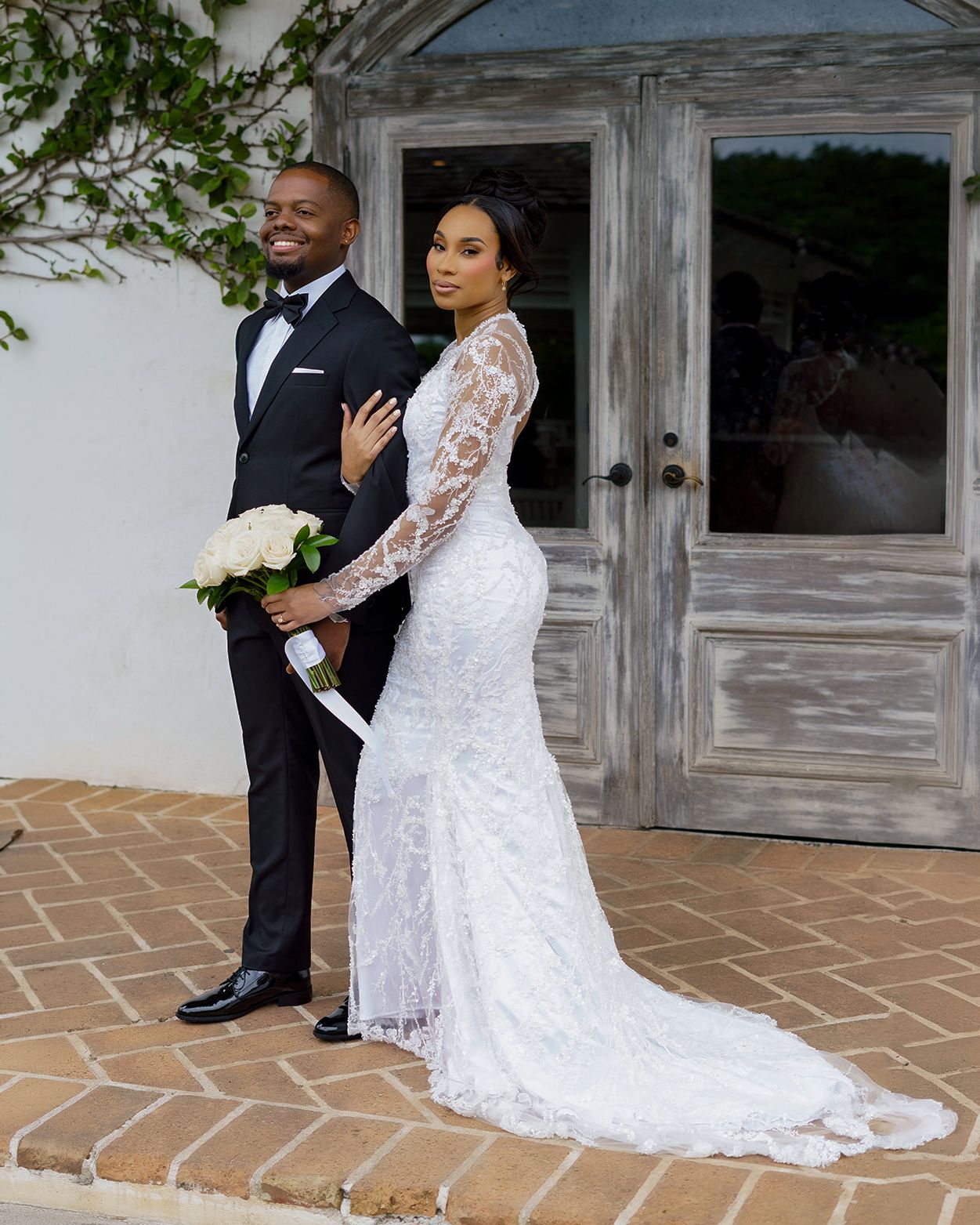 A bride in a lace mermaid gown and groom in a black tuxedo pose elegantly outside a rustic venue.