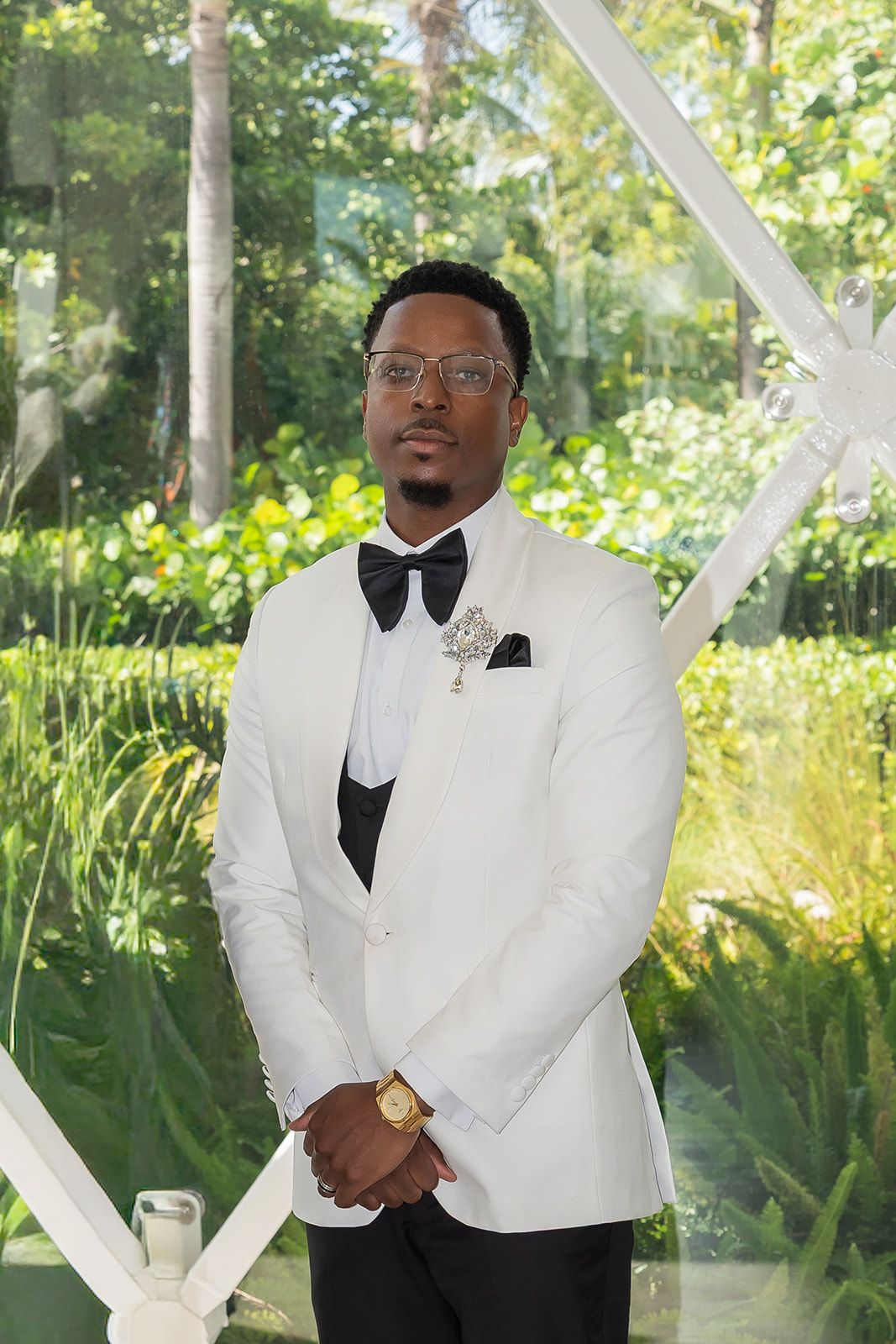 A well-dressed Black man in a white tuxedo jacket and black bow tie stands outdoors amid lush green foliage.
