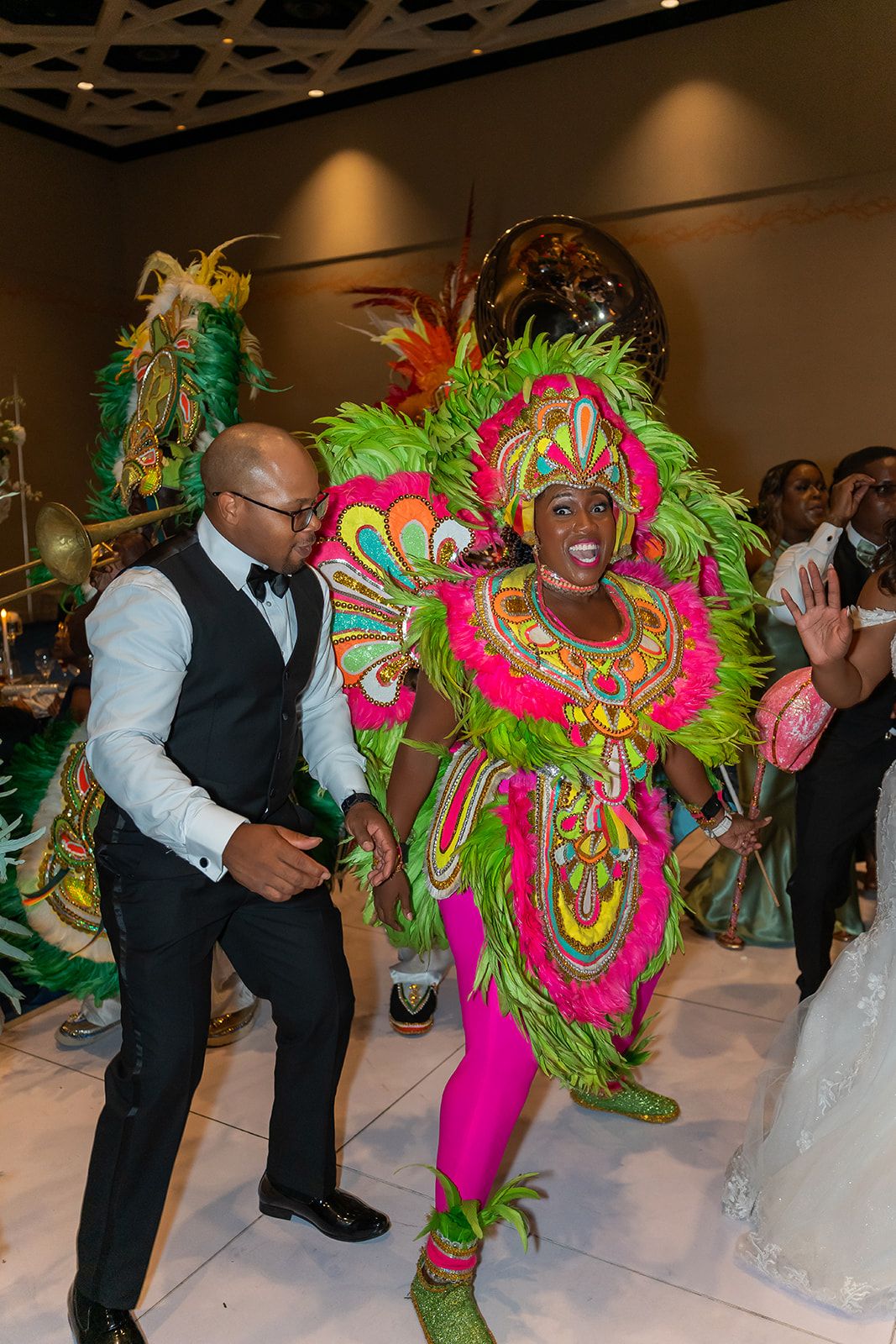 Vibrant carnival dancer in colorful feathered costume performs with a man in a tuxedo vest at an indoor celebration.