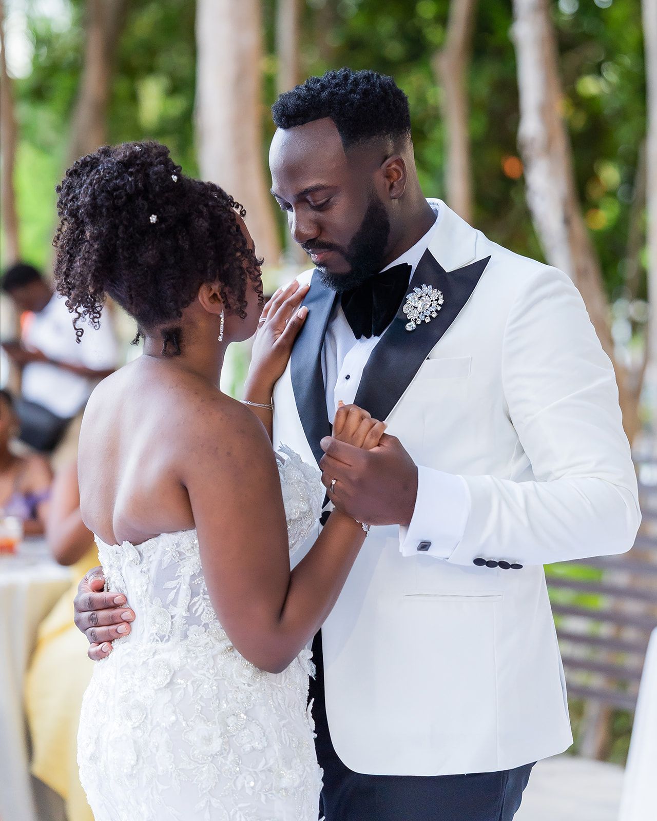 Groom in white tuxedo gazes at bride during outdoor first dance, bride touching his face tenderly at wedding.