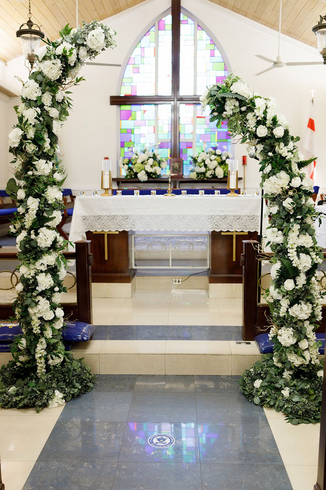 Church altar adorned with white floral arch and greenery for a wedding ceremony, with stained glass window backdrop.