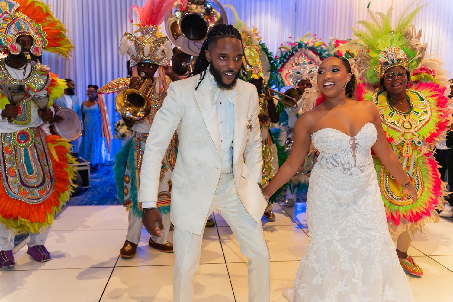 Bride and groom dancing at Caribbean-themed wedding reception with colorful costumed performers in vibrant feathered outfits.