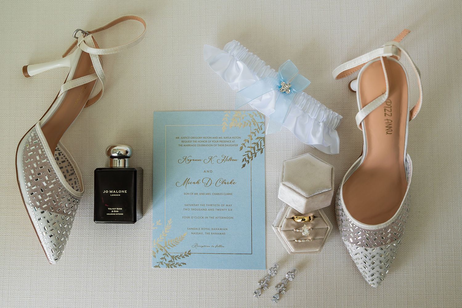 Bridal flat shoes with crystals, wedding invitation, perfume, garter, ring box, and earrings arranged on white surface.