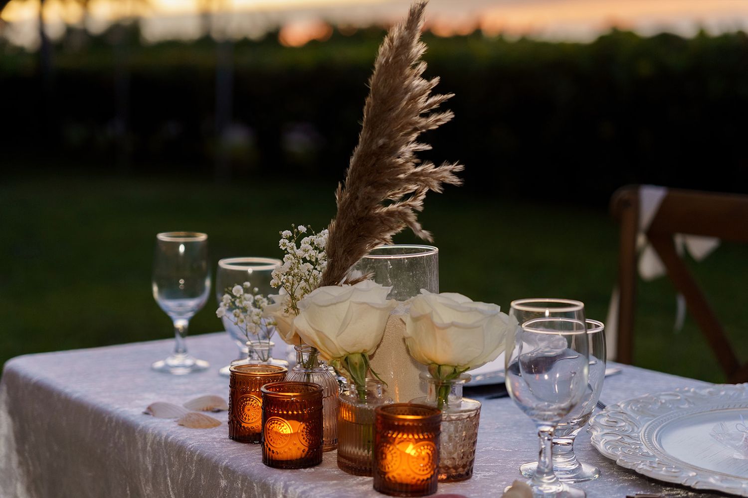 Outdoor wedding reception table with white roses, pampas grass, amber votives at sunset.