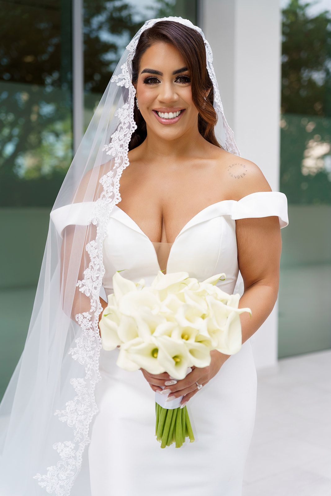 Smiling bride in off-shoulder white gown holds calla lily bouquet, wearing lace-trimmed veil for elegant wedding portrait.