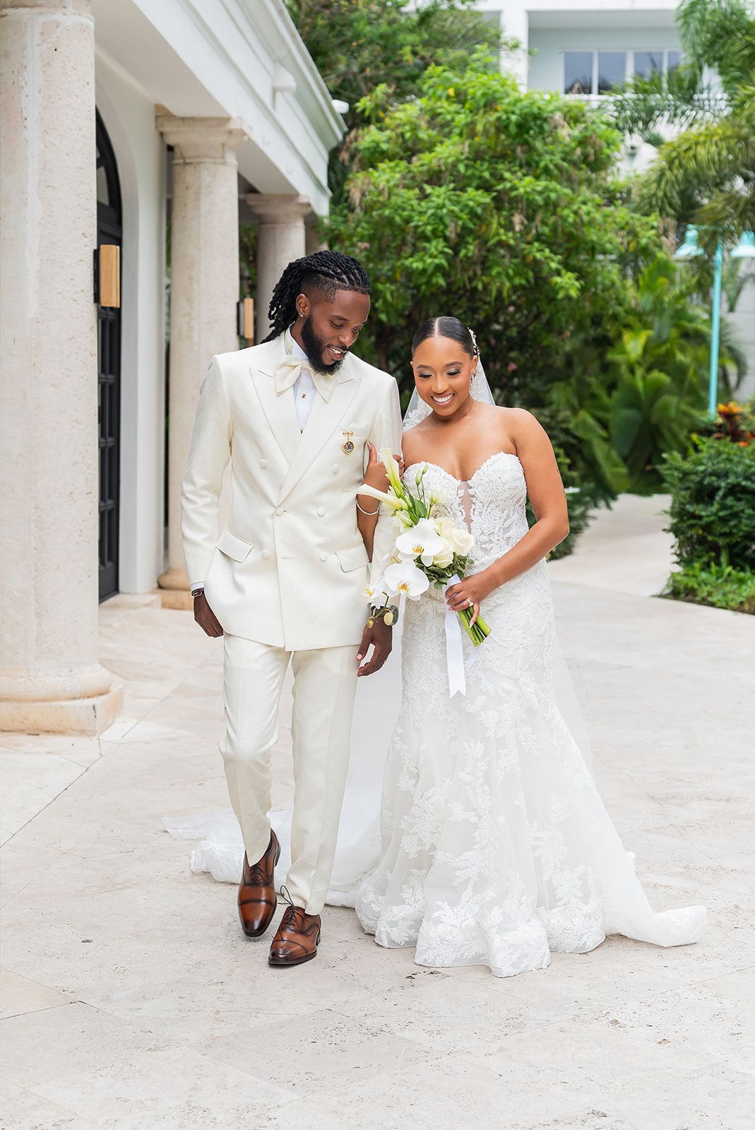 Newlywed couple walking together outdoors, groom in cream suit, bride in lace gown holding white bouquet.