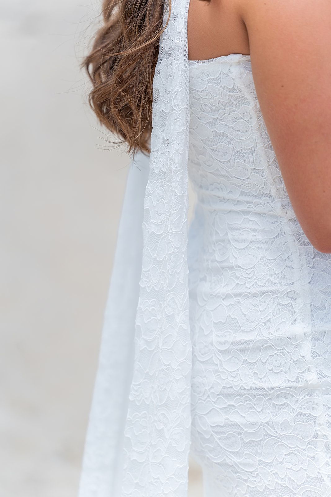 Close-up of a bride in a white lace halter wedding dress with a flowing chiffon panel at a beach.