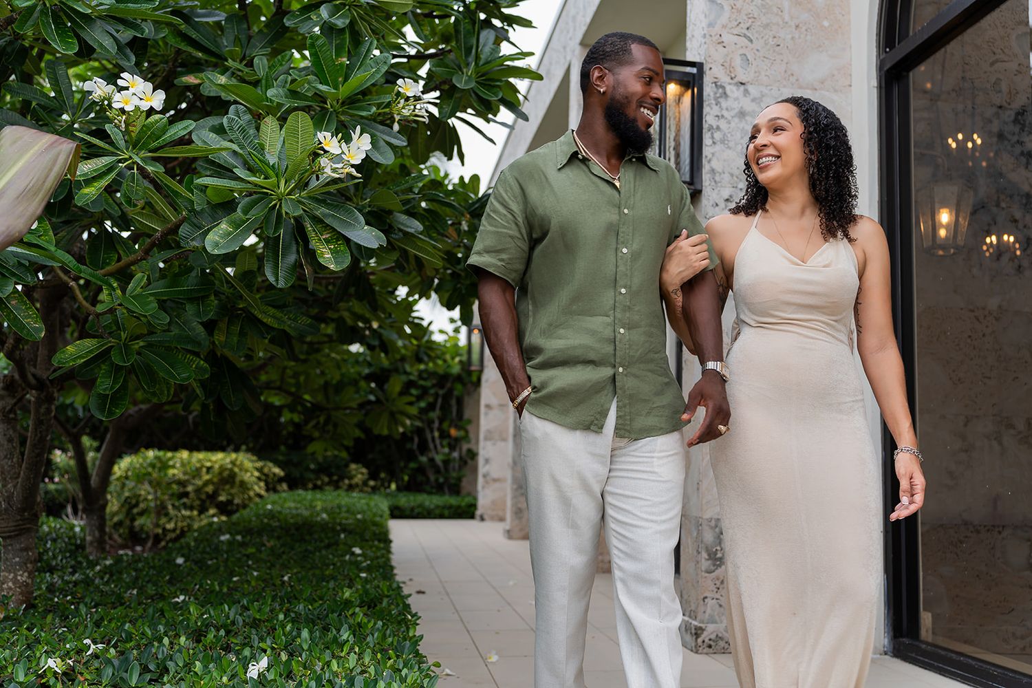 A smiling pregnant woman in a beige dress walks arm-in-arm with a man in green shirt outdoors near tropical flowers.