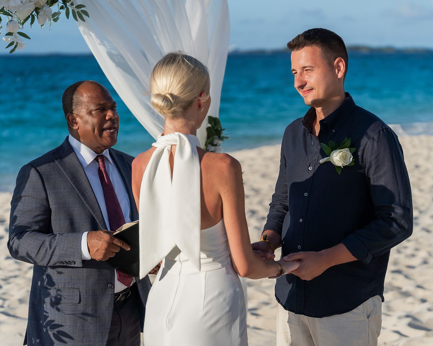 Couple exchanging wedding vows on a tropical beach with an officiant, ocean backdrop, and white fabric arch.