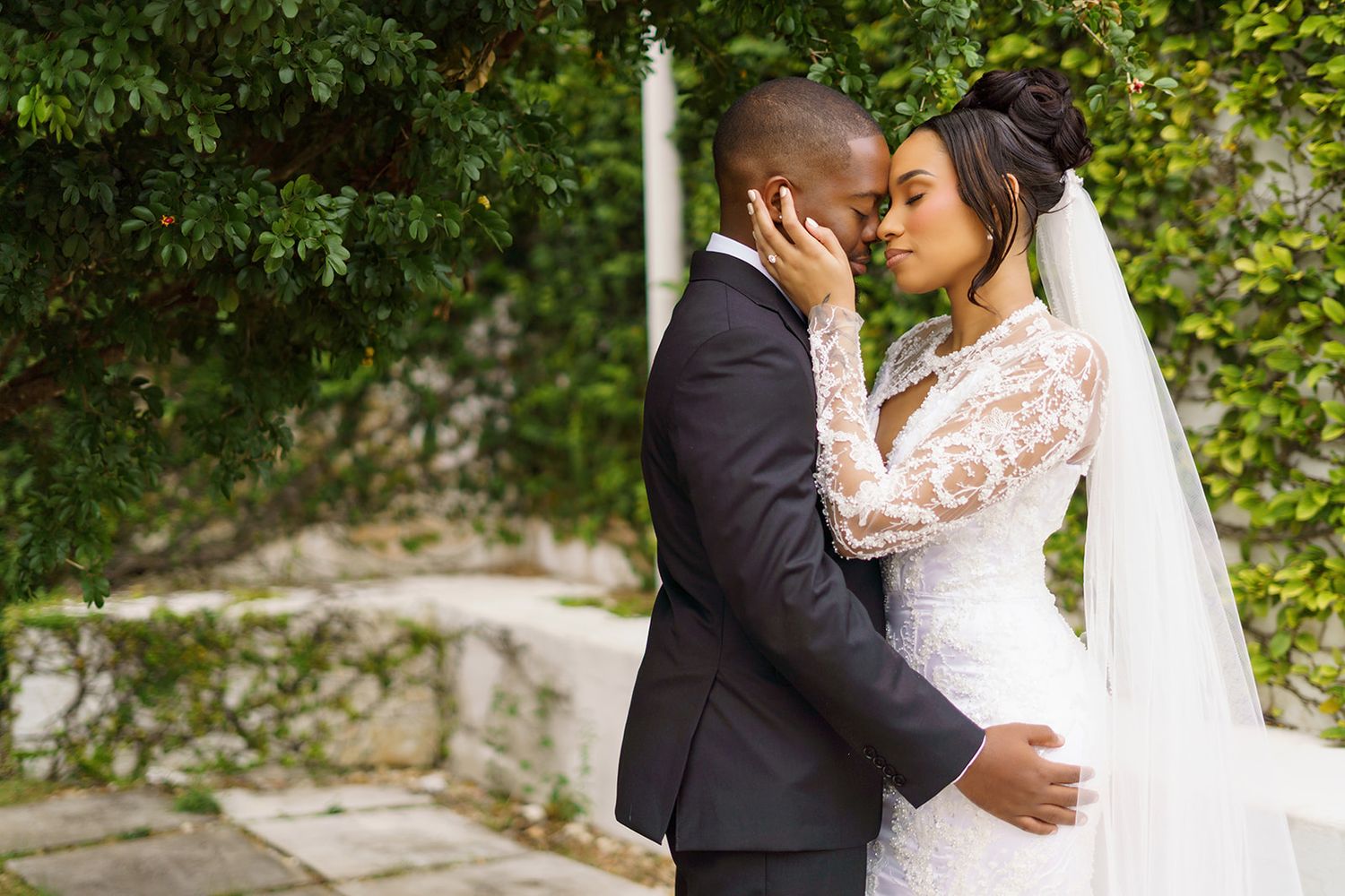 Newlyweds in wedding attire share an intimate moment surrounded by lush green foliage.