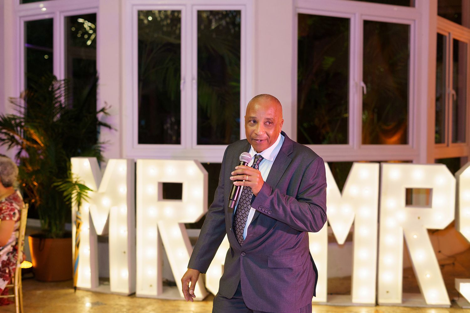 A man in a suit speaks into a microphone at a wedding reception in front of lit MR & MRS letters.