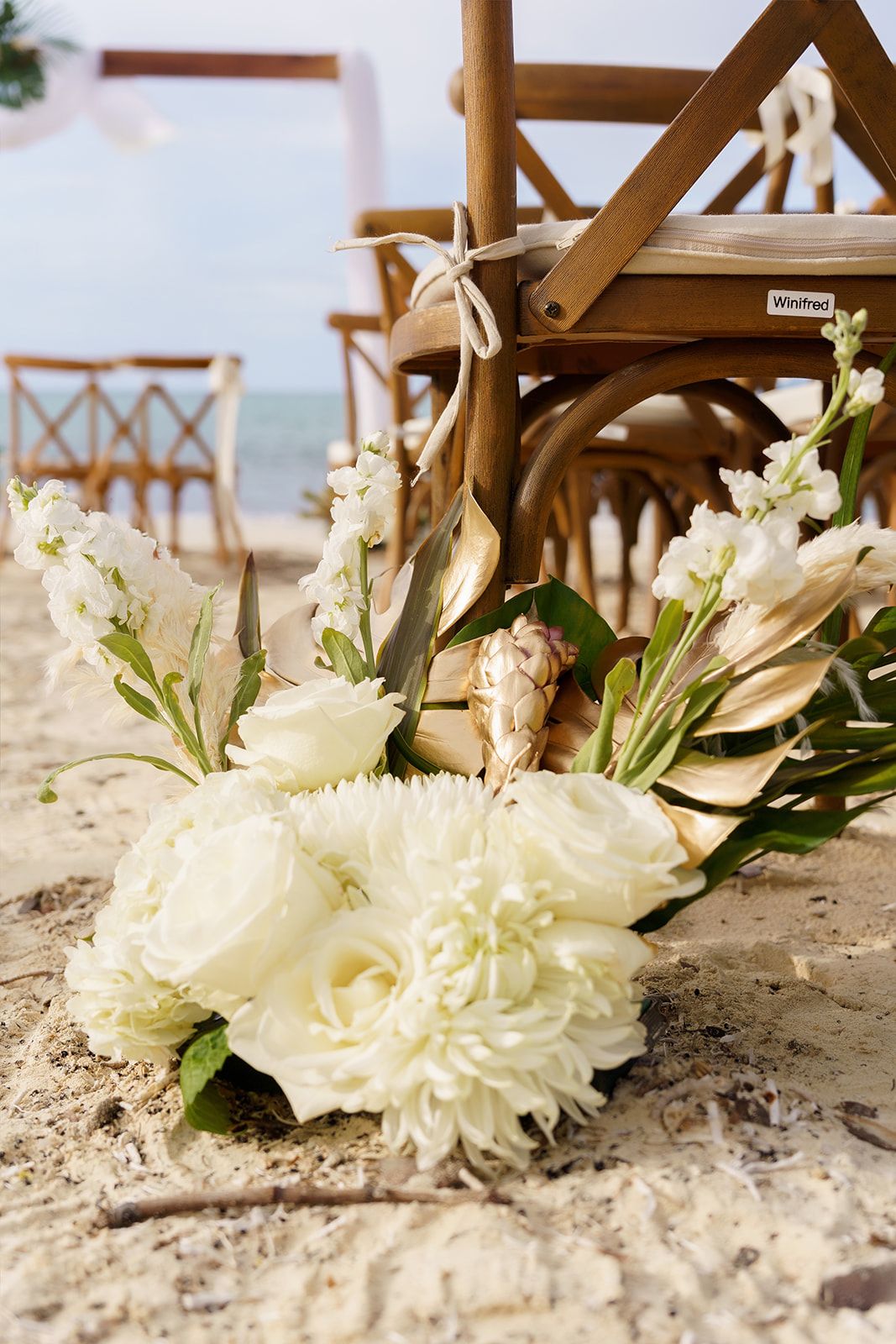 White floral bouquet with roses and dahlias on sandy beach beside wooden chairs at an elegant outdoor wedding ceremony.