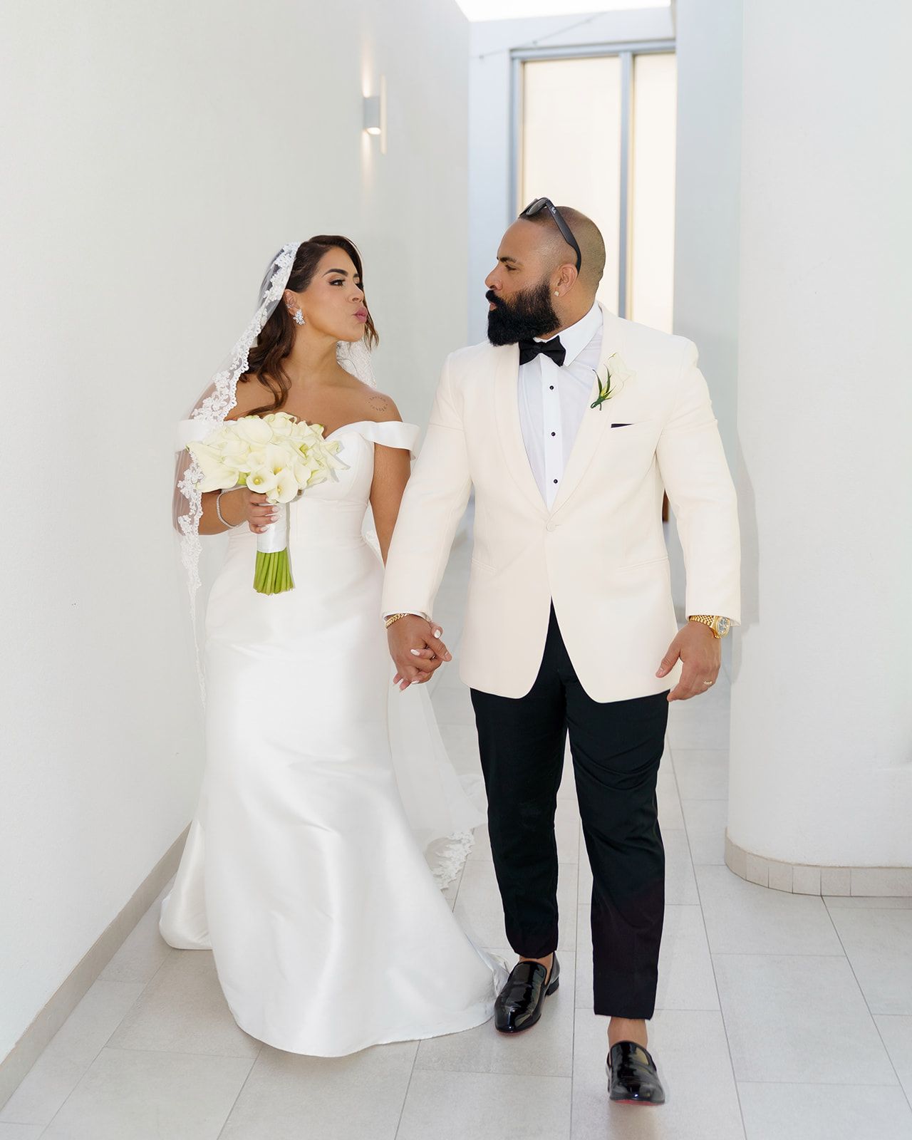 Bride in off-shoulder white gown holding bouquet walks with groom in cream tuxedo and black pants in bright hallway.