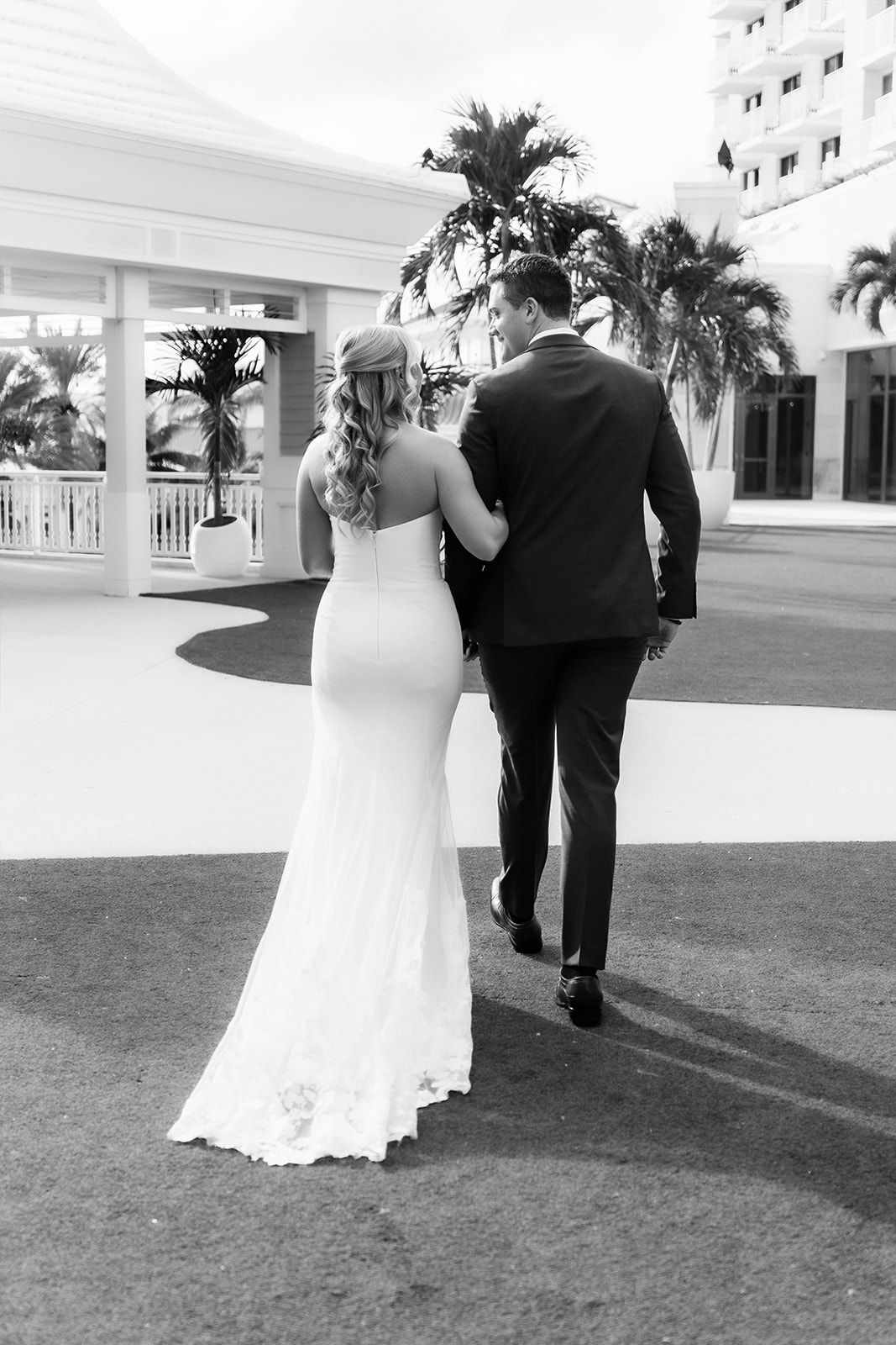 Black and white photo of a bride and groom walking away, showcasing her fitted lace gown near a tropical venue.