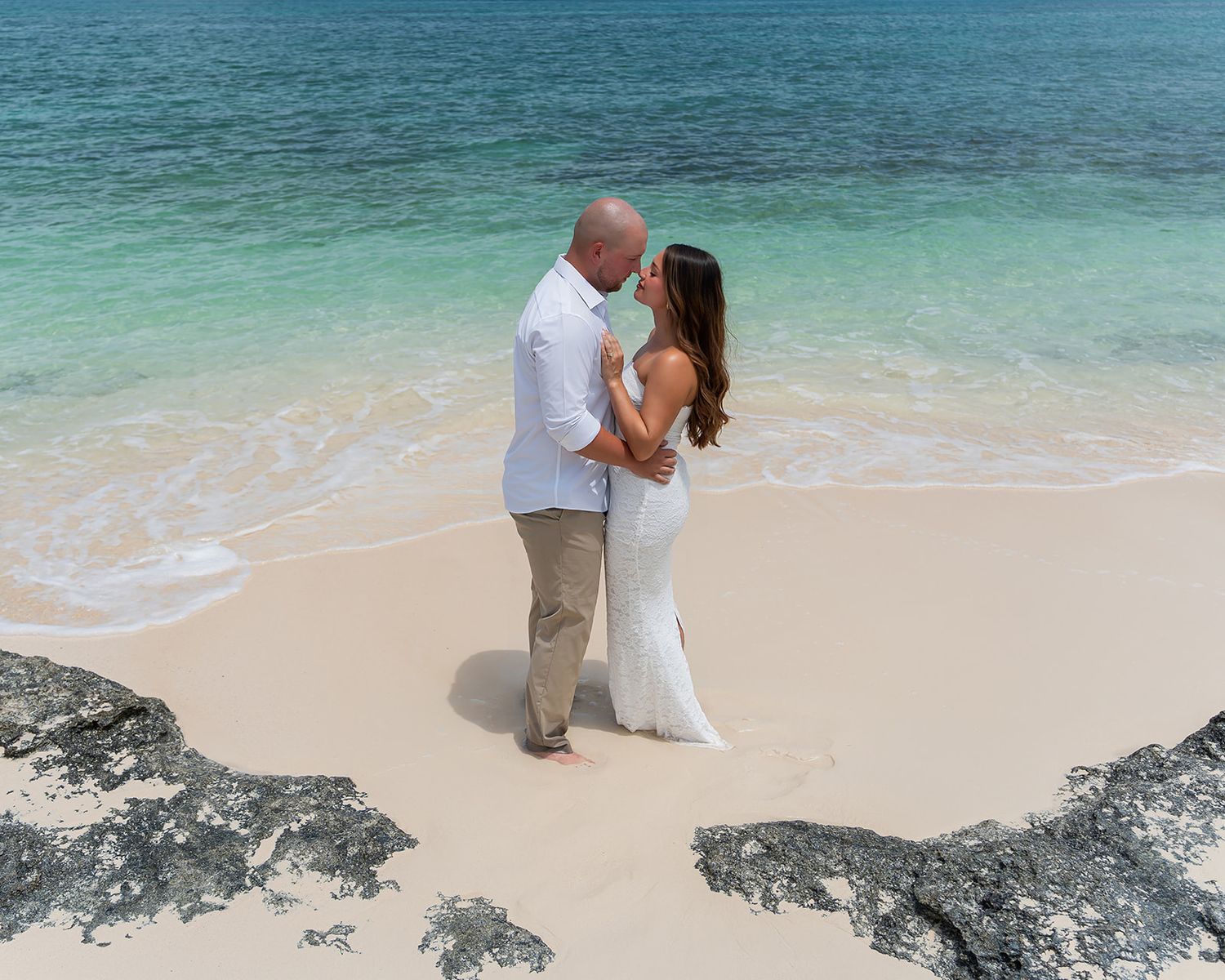 Couple sharing a romantic kiss on a white sand tropical beach with turquoise water, dressed in wedding attire.