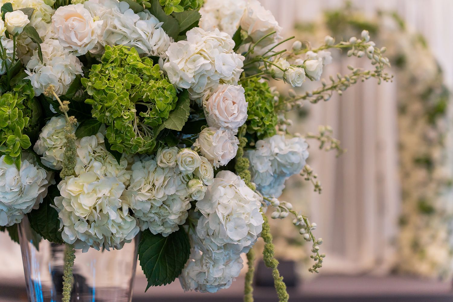 Lush wedding floral centerpiece featuring white hydrangeas, green viburnum, and soft blue blooms in a glass vase.