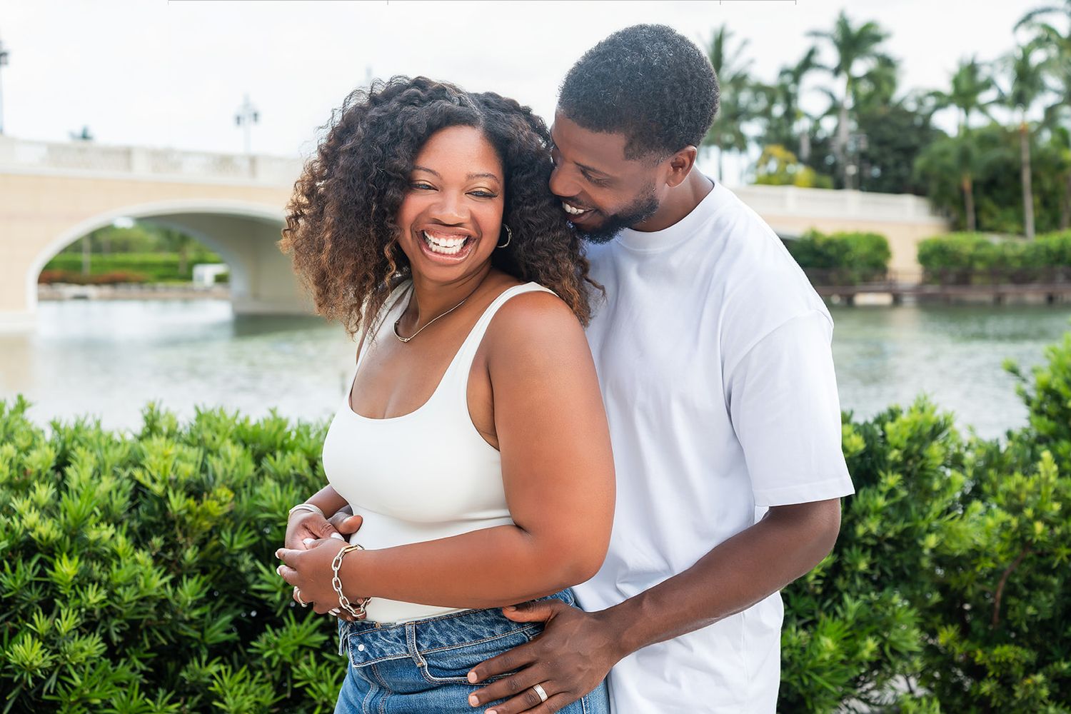 A joyful Black couple embraces outdoors near a bridge and lush greenery, both wearing white tops and smiling.