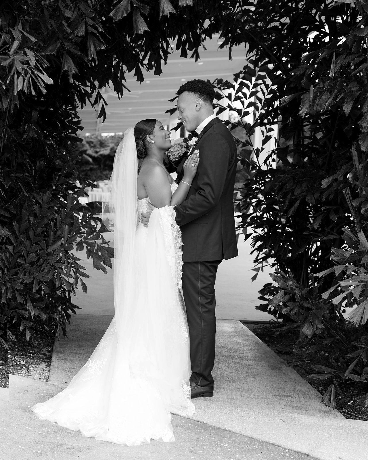 Black and white photo of a bride and groom sharing a romantic moment under a lush floral arch at their wedding.