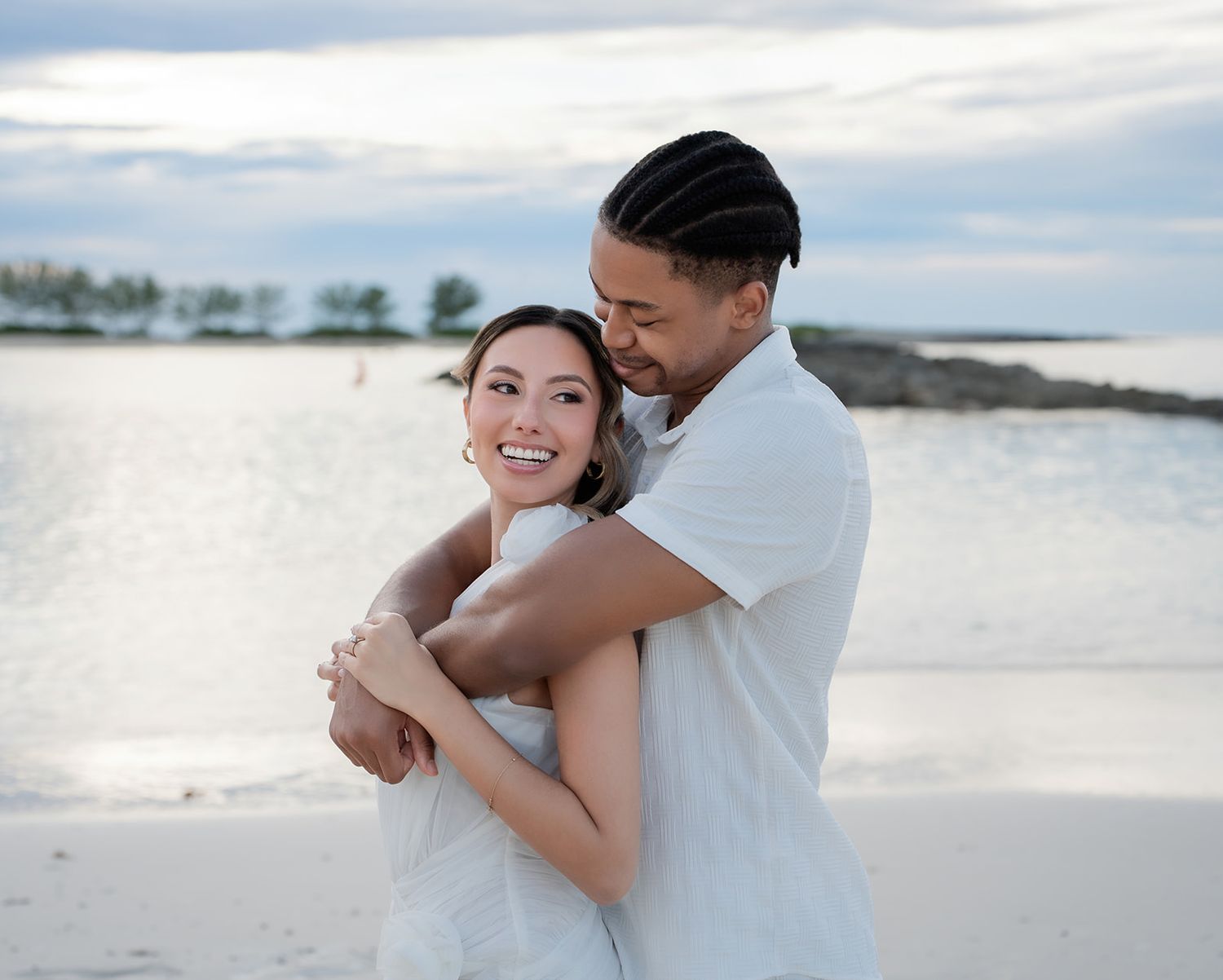 Couple embracing and smiling on a sandy beach at sunset, wearing white outfits with calm ocean water in the background.