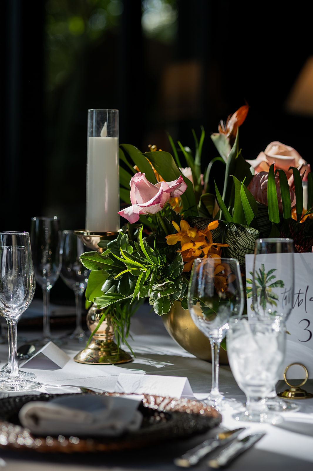 Elegant wedding table setting with pink roses, tropical greenery, gold candlestick, crystal glasses, and a table number card.