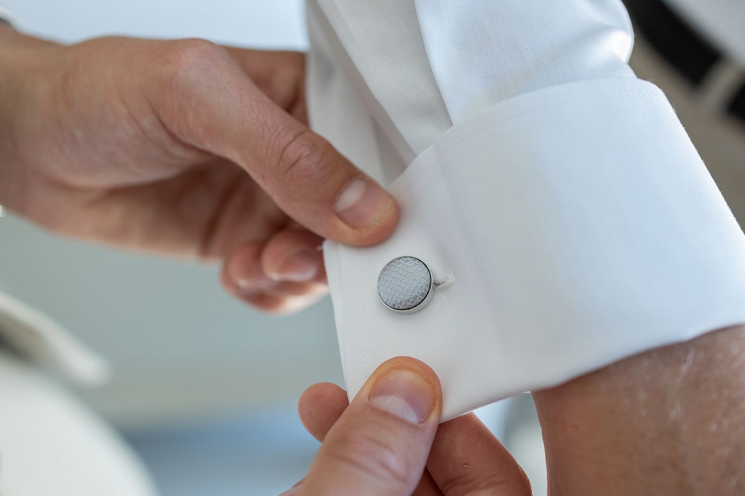 Close-up of hands fastening a silver cufflink on a white dress shirt sleeve.