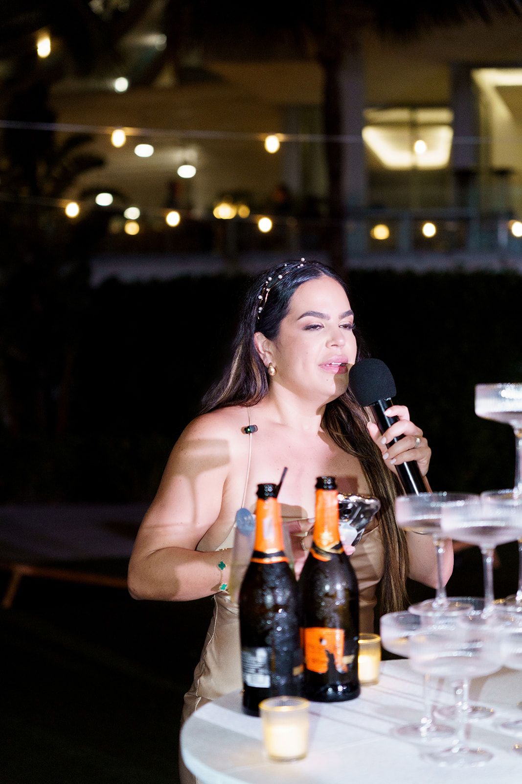 Woman speaking into microphone at outdoor evening party with champagne tower and bottles on table.