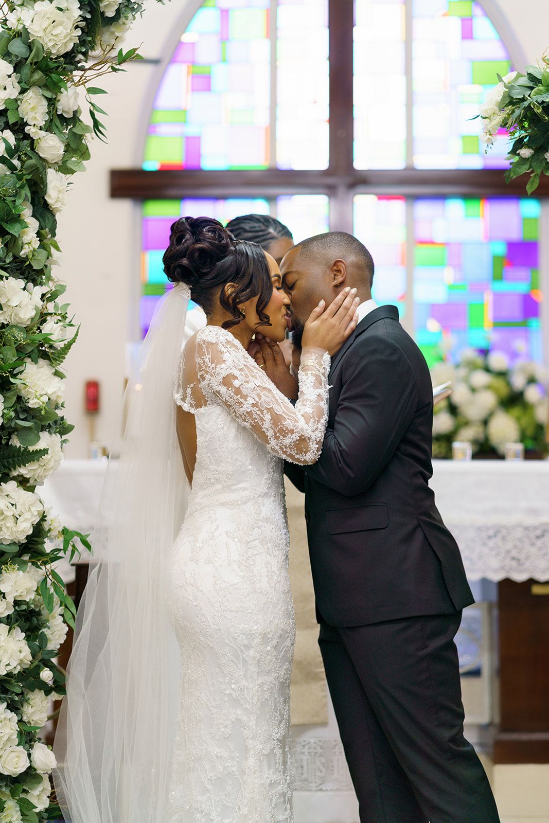 A bride and groom share their first kiss at the altar in a church with a colorful stained glass window backdrop.