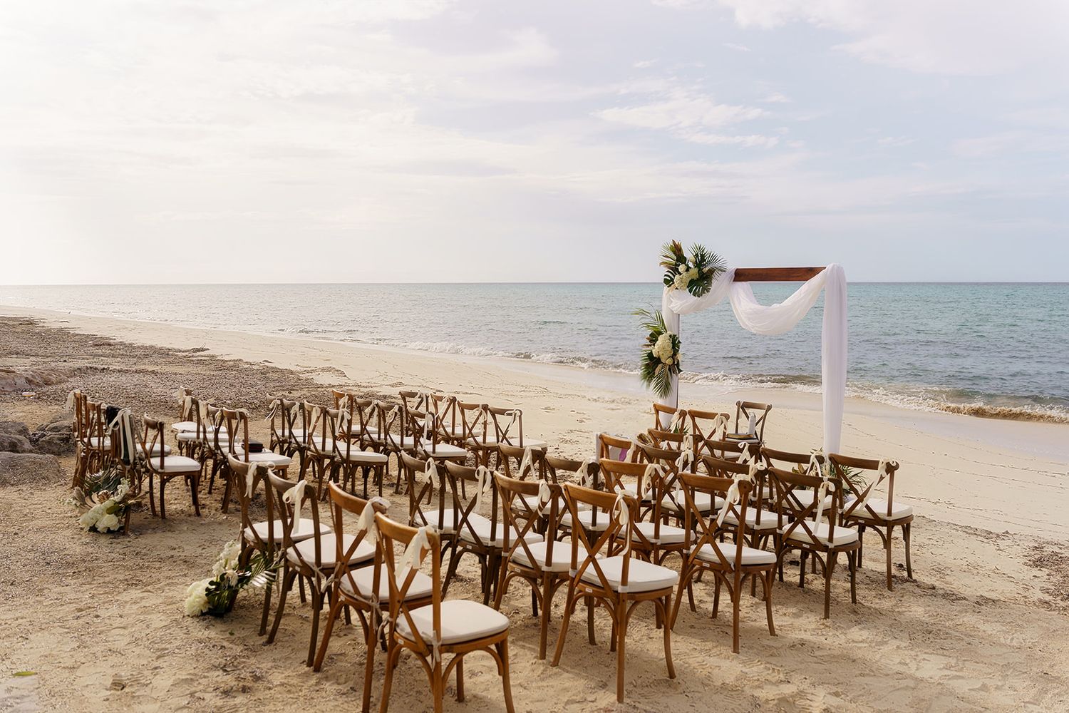 Beach wedding ceremony setup with wooden cross-back chairs and white floral arch by the ocean.