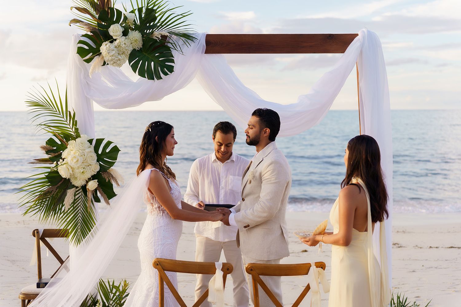 Couple exchanging vows at elegant beach wedding altar adorned with white draping and tropical floral arrangements.