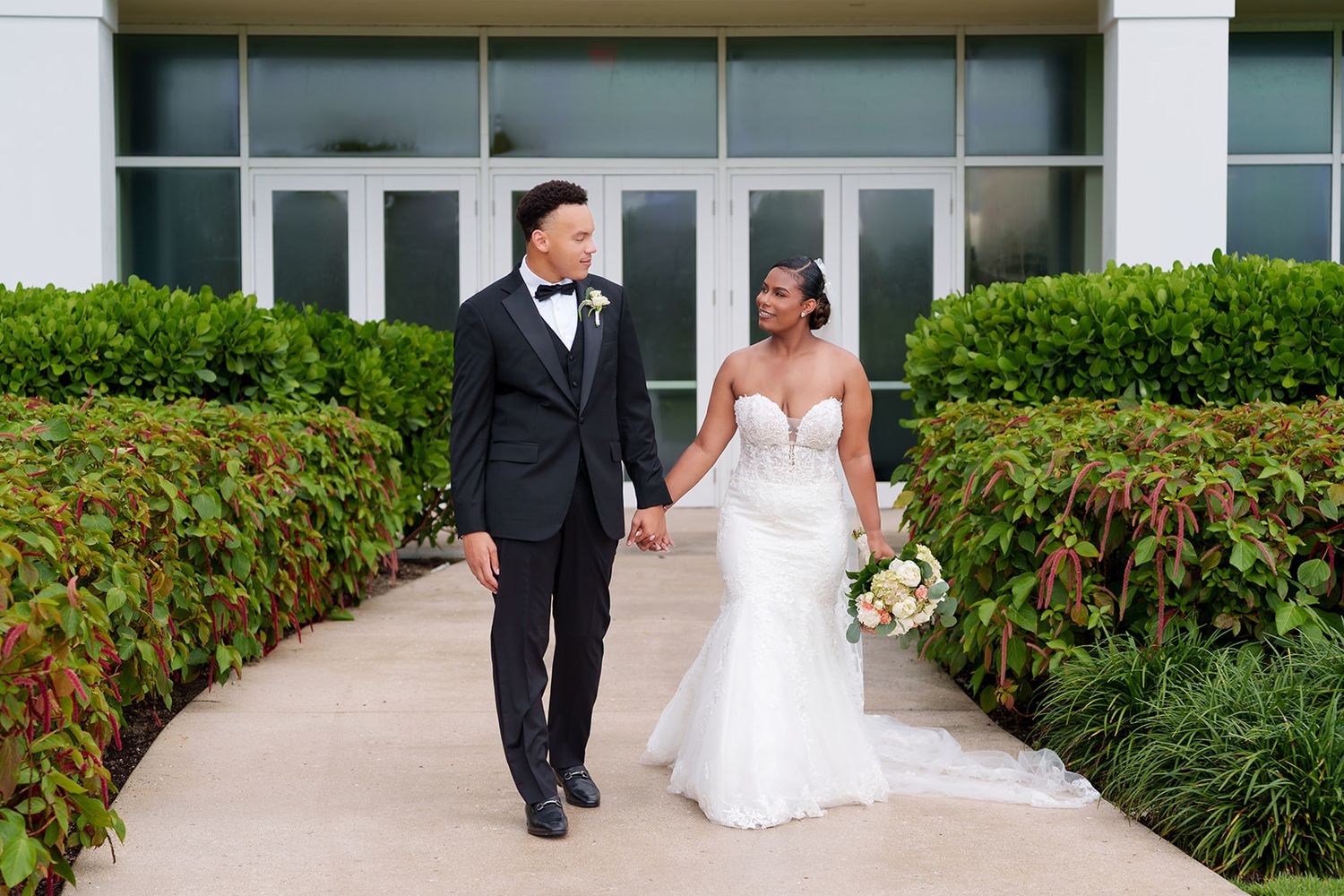 Bride and groom walk hand in hand along a garden pathway, she in a strapless gown, he in a black tuxedo.