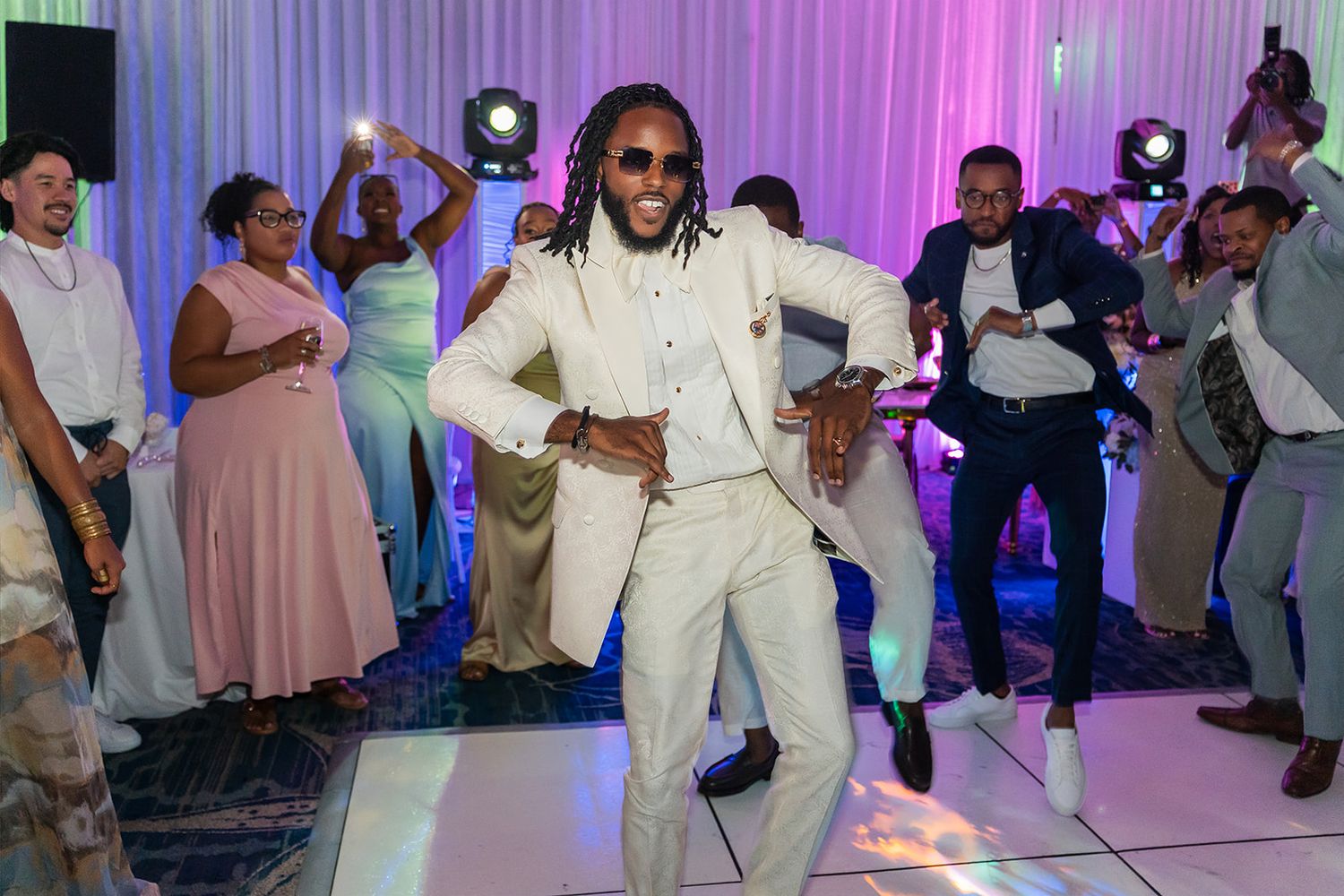 Man in white suit dancing joyfully at a wedding reception with guests celebrating in the background.