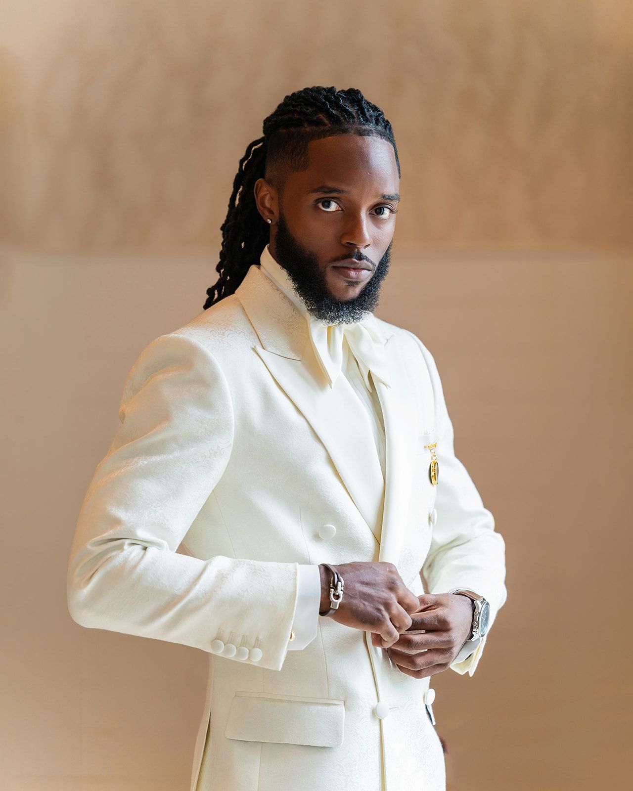 Man in cream white suit with braided hair and beard posing confidently.