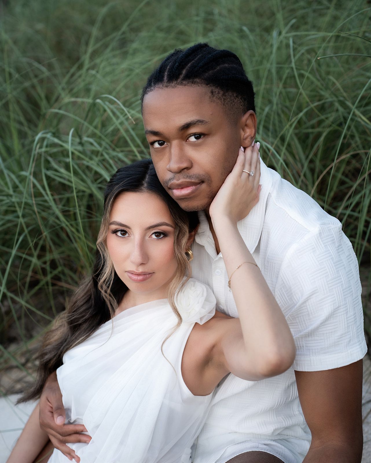 Romantic couple in matching white outfits posing together among tall green beach grass in a professional photo.