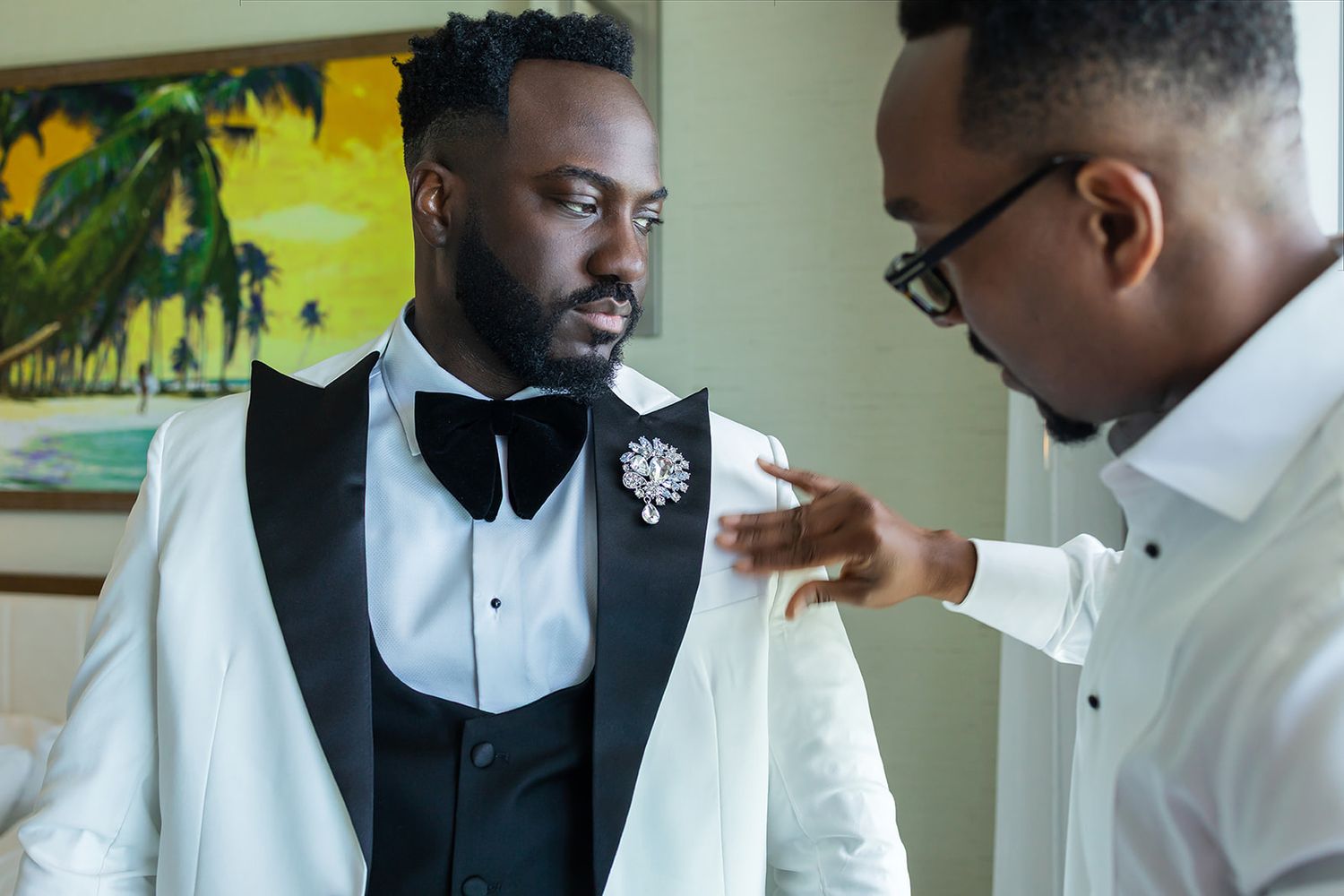 A groom in a white tuxedo with bow tie and brooch being assisted by a man adjusting his jacket lapel.