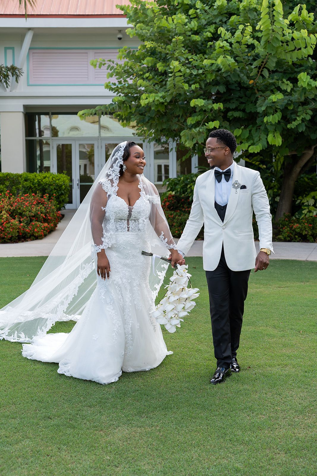 Bride and groom walk hand-in-hand on green lawn, she in lace mermaid gown with veil, he in white tuxedo jacket.