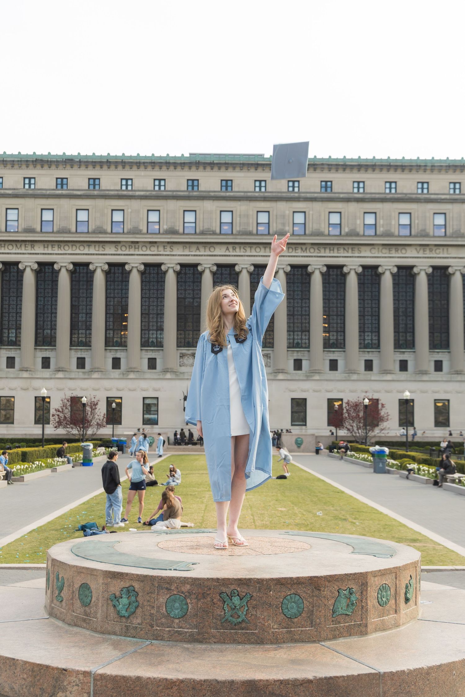 Young woman in blue graduation gown standing on fountain pedestal, holding diploma aloft in front of Columbia University.