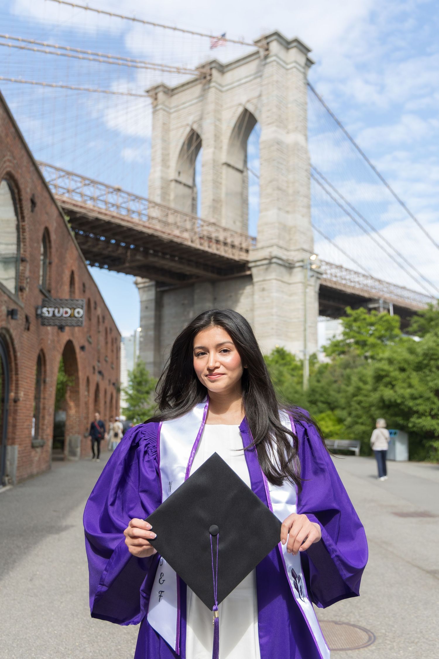 Graduate in purple cap and gown poses proudly in front of the Brooklyn Bridge on a sunny day.