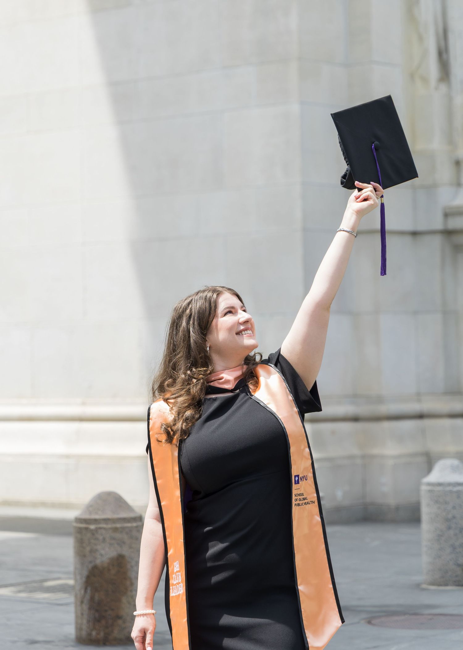 A smiling graduate in black dress and orange sash raises her mortarboard cap outdoors near a stone building.