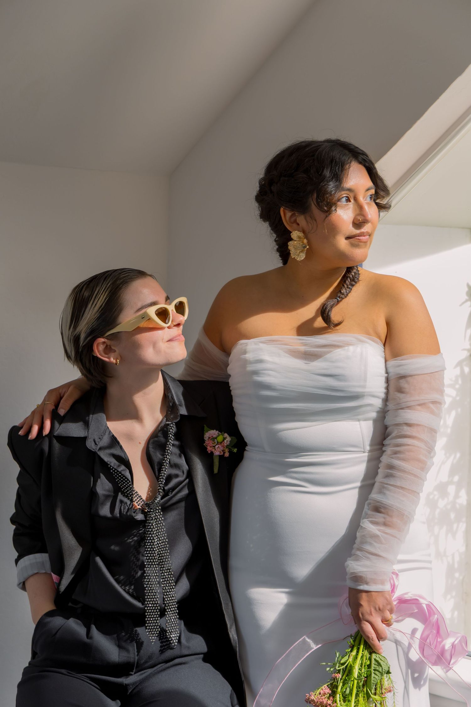 Bride and groom pose together in formal attire against a bright window backdrop.