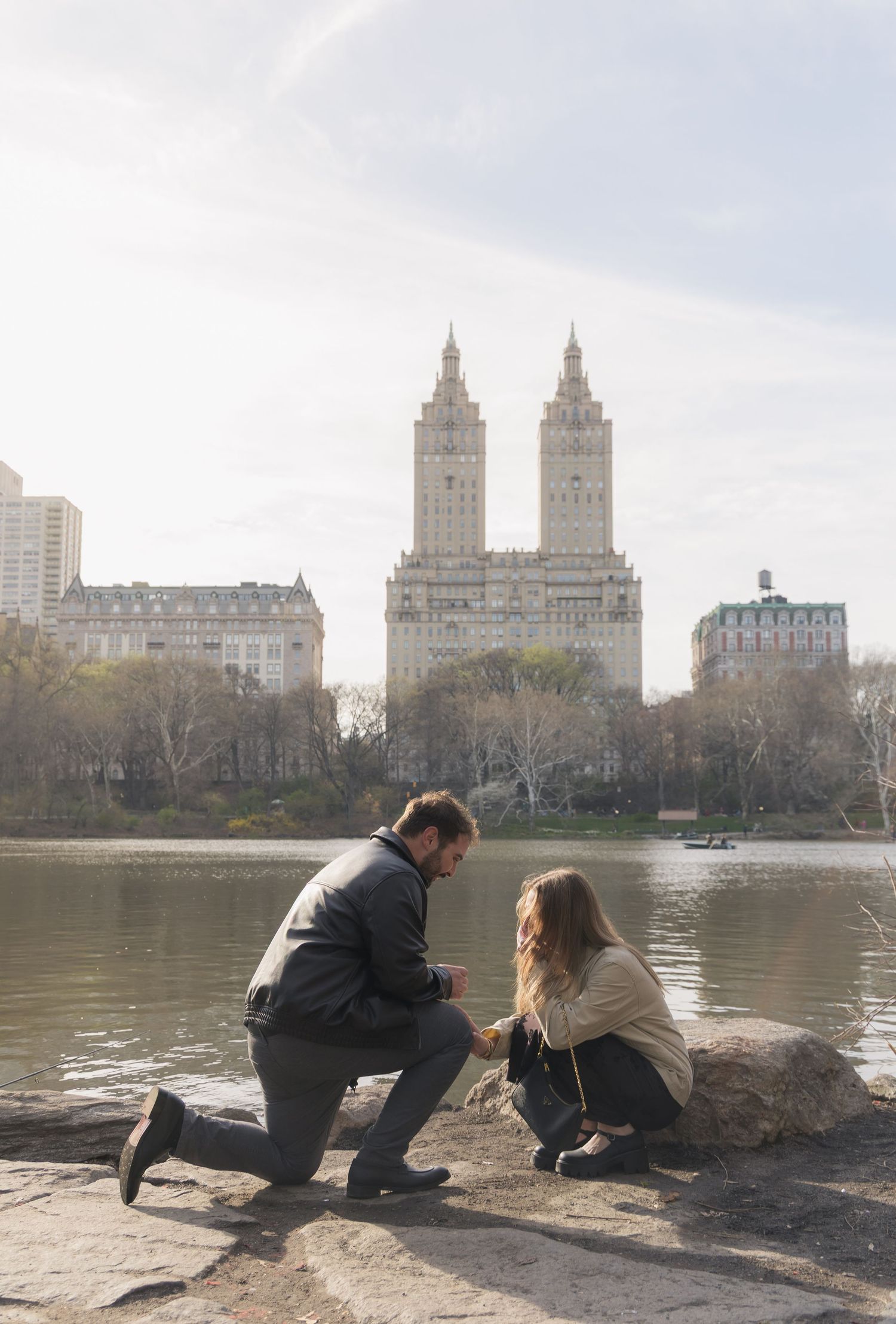 Man proposes on one knee to woman by Central Park lake with iconic San Remo building in NYC background.