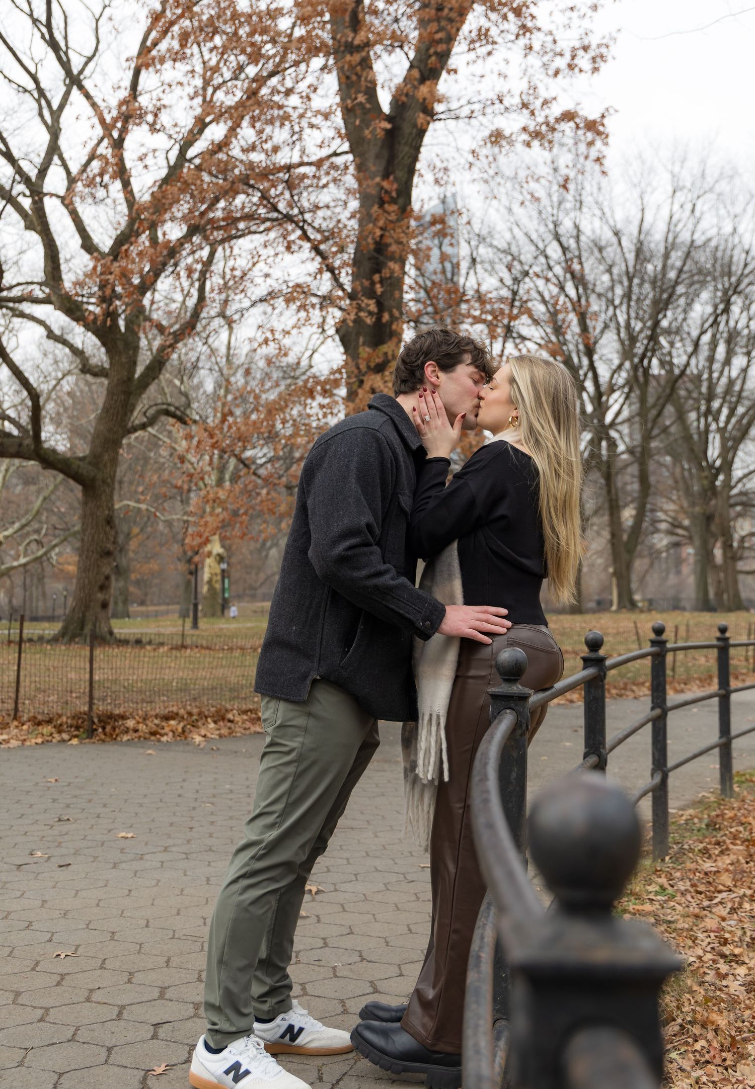 Couple sharing a romantic kiss in Central Park during autumn, surrounded by bare trees and fallen leaves.