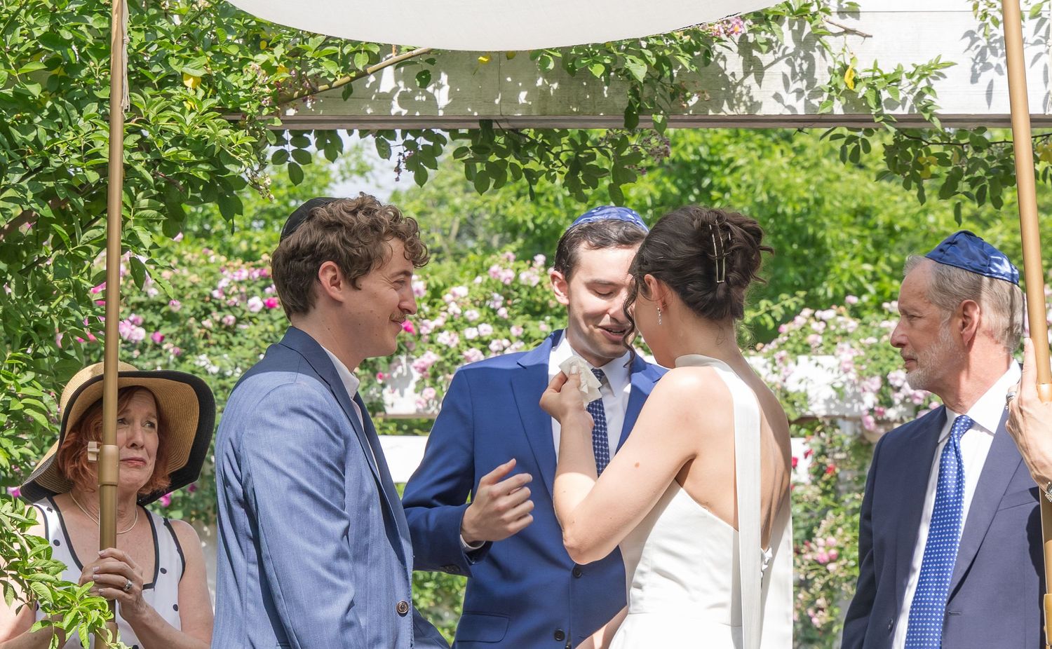 Outdoor wedding ceremony with bride and groom sharing a moment under a floral chuppah surrounded by guests.