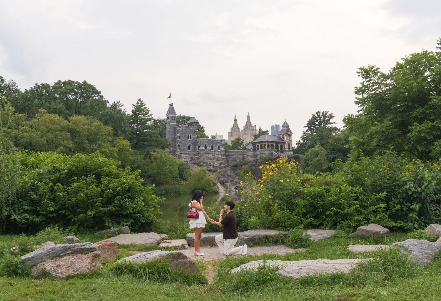 Couple holding hands on rocks before Belvedere Castle surrounded by lush greenery in Central Park, NYC.
