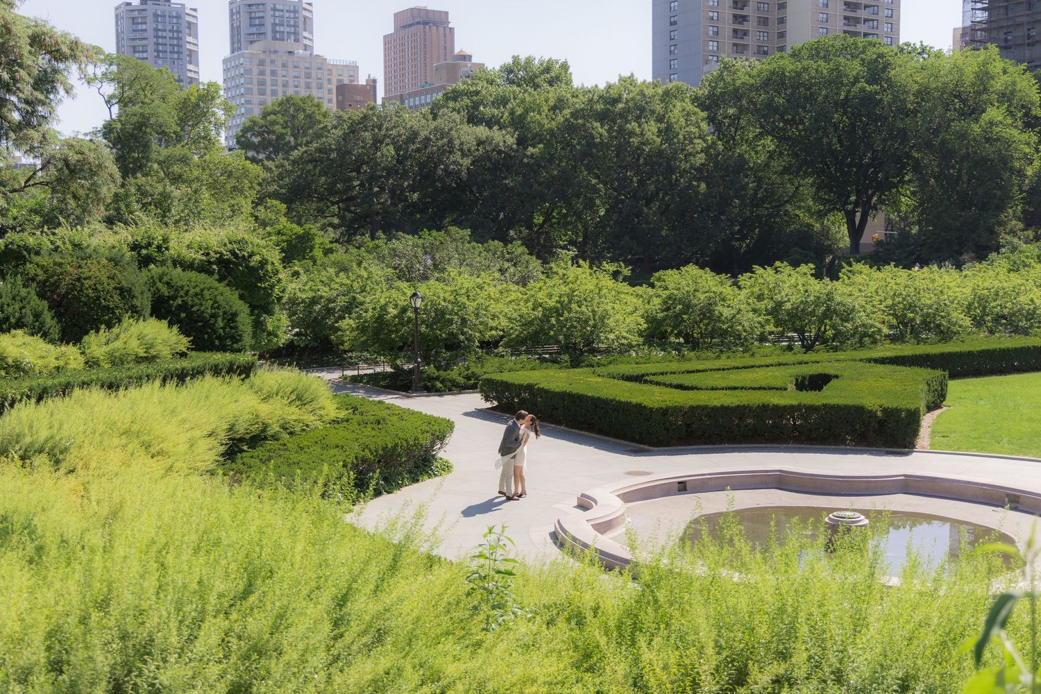 Person strolling through lush Central Park garden with manicured hedges, green trees, and NYC skyline in background.