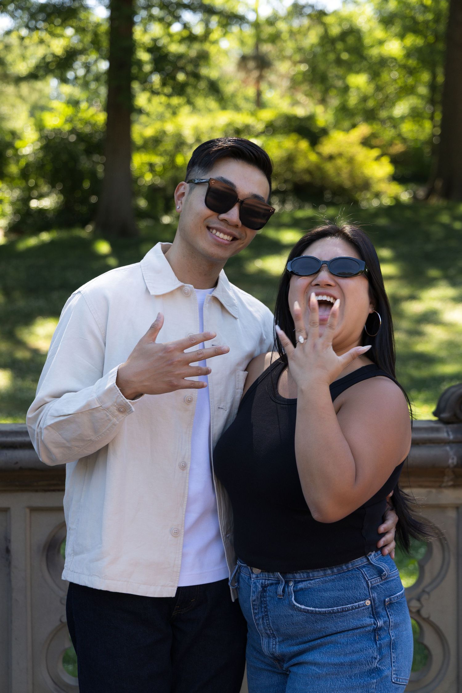 Happy couple in sunglasses showing off engagement ring outdoors in a park setting.