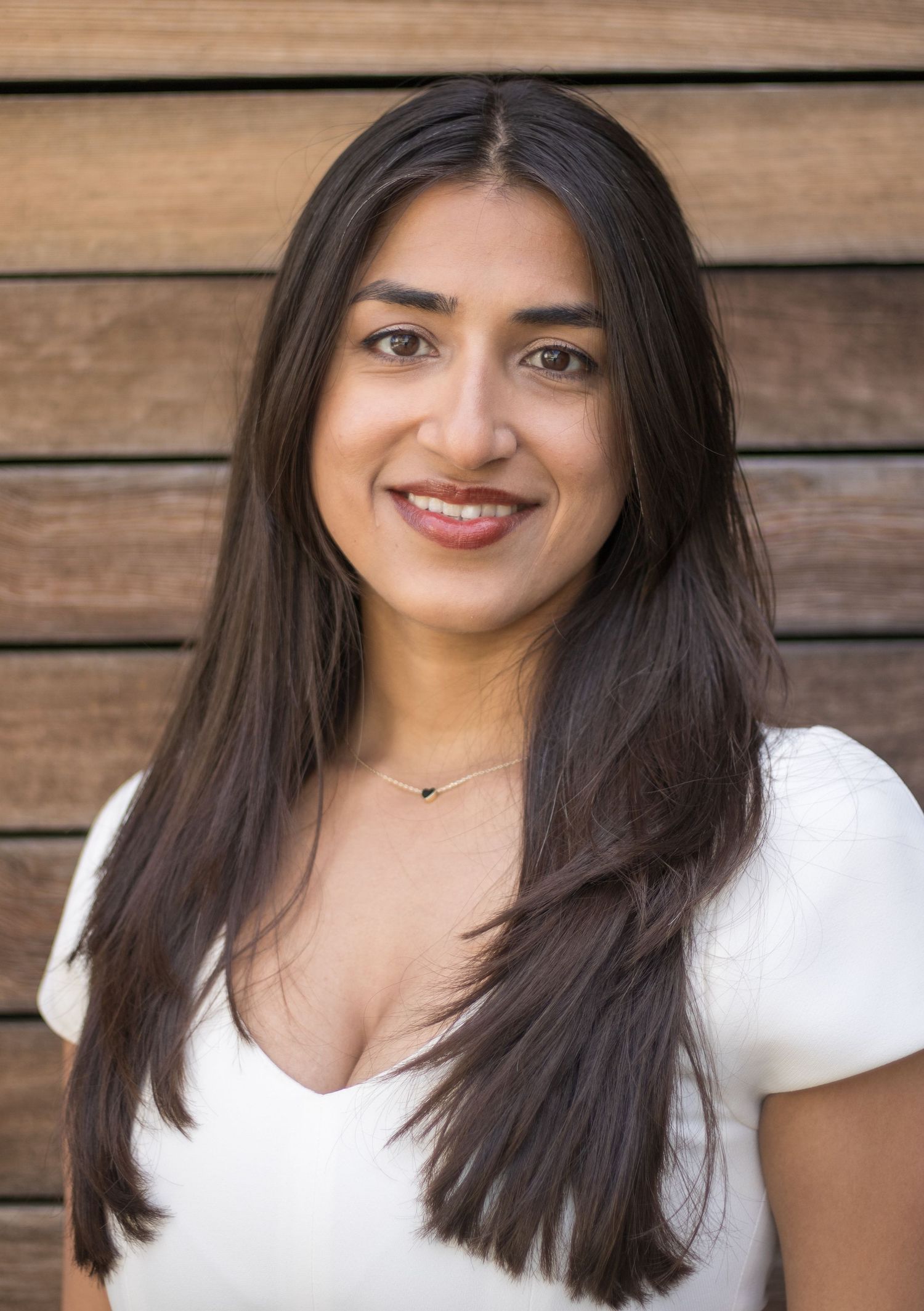 Professional headshot of a person with long dark hair wearing a white top against a wooden wall background.