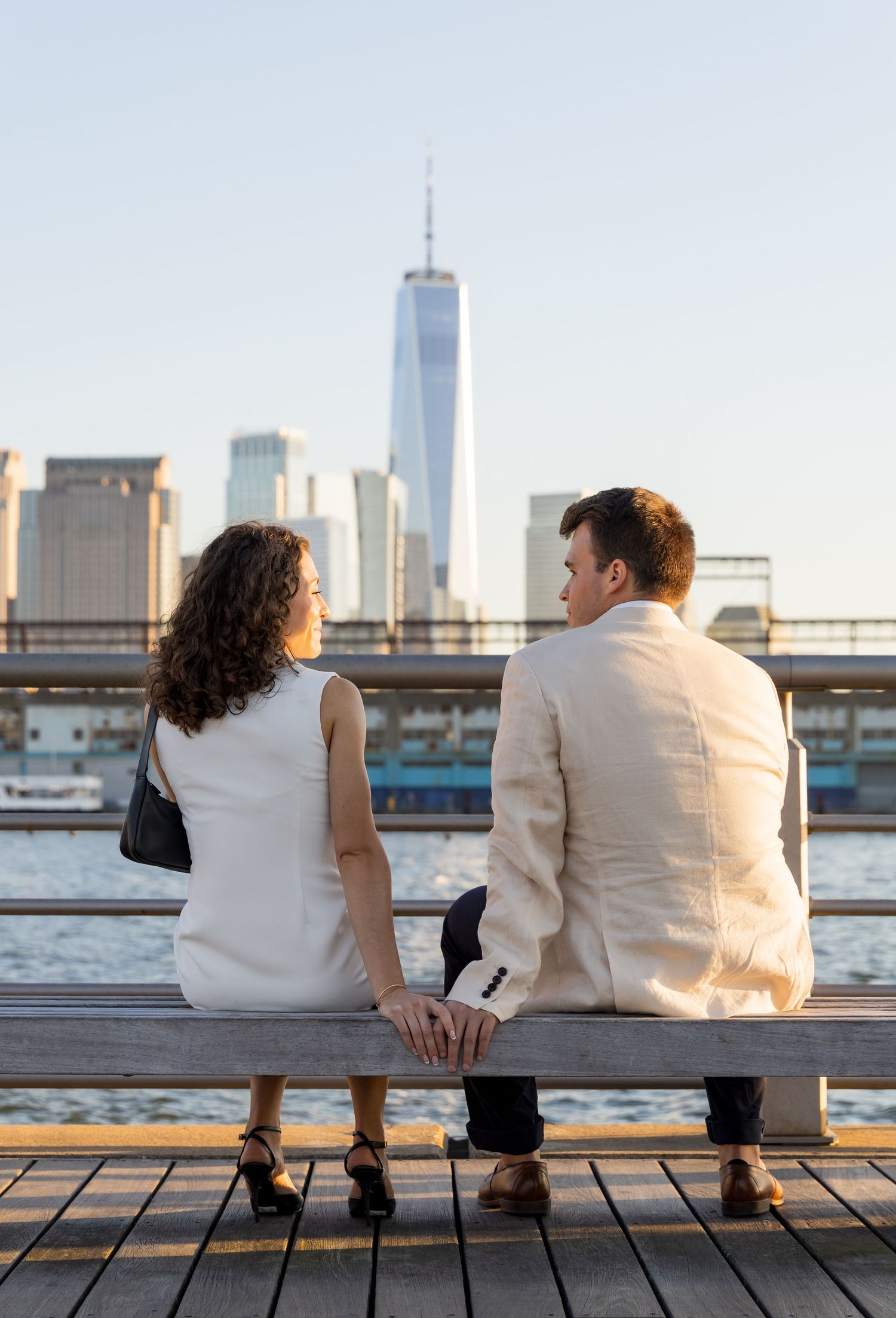 Couple sitting on a bench holding hands with One World Trade Center and NYC skyline in the background.